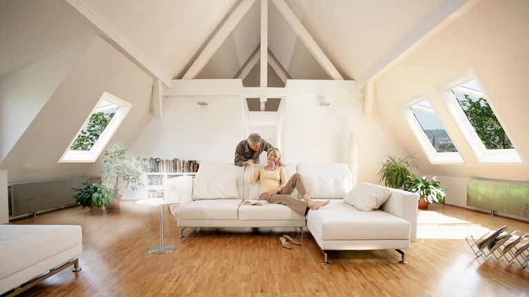 ein Paar wird in seiner Dachwohnung im Wohnzimmer gezeigt, die Dachfenster an den Seiten sind geöffnet zum Lüften